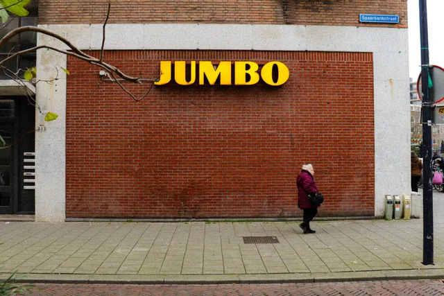 Elderly woman walking by Jumbo supermarket with red brick wall background in urban setting. Winkels in de wijk: hoe u het lokale aanbod vindt en vergelijkt